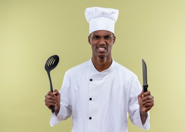 Joven cocinero afroamericano molesto en uniforme de chef sostiene cuchillo y espátula aislado sobre fondo verde con espacio de copia