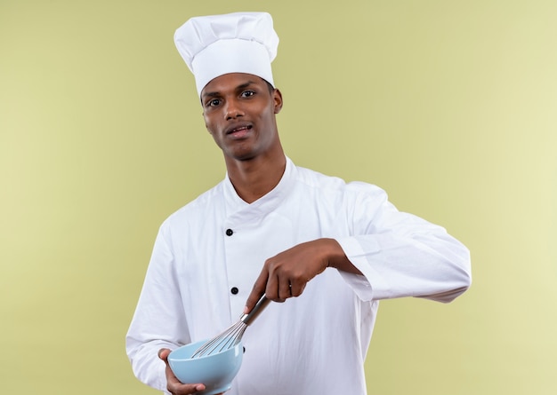 Joven cocinero afroamericano molesto en uniforme de chef sostiene un cuenco y batidor aislado sobre fondo verde con espacio de copia