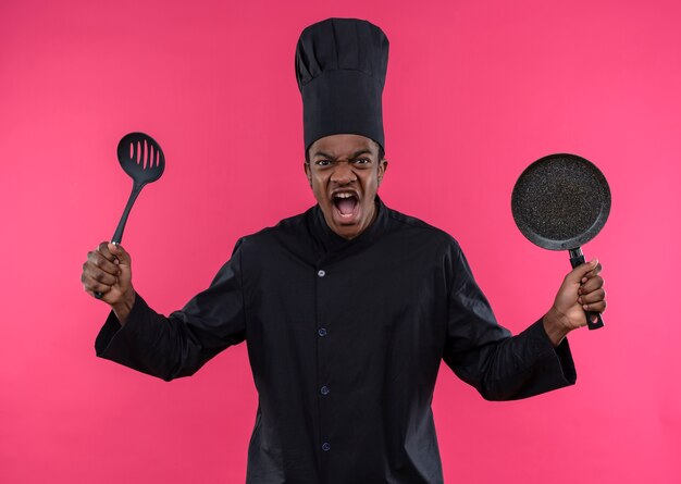 Joven cocinero afroamericano molesto en uniforme de chef sostiene una sartén y una espátula aisladas sobre fondo rosa con espacio de copia