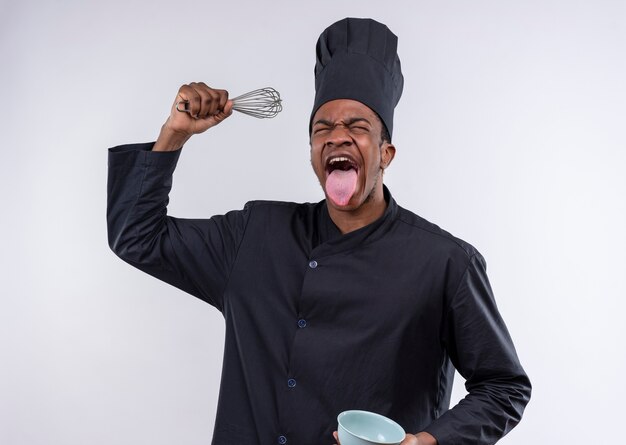 Joven cocinero afroamericano molesto en uniforme de chef sostiene un tazón y un batidor de lengua pegado aislado sobre fondo blanco con espacio de copia