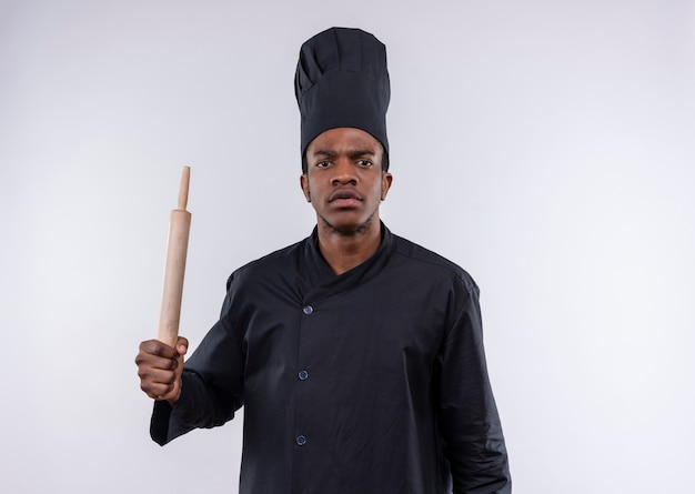 Joven cocinero afroamericano molesto en uniforme de chef tiene rodillo aislado sobre fondo blanco con espacio de copia