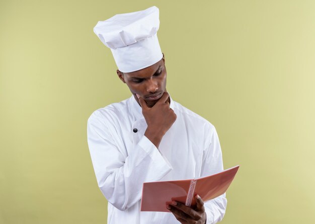 Joven cocinero afroamericano pensativo en uniforme de chef mira portátil aislado en la pared verde