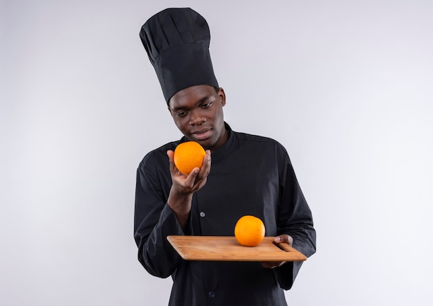 Joven cocinero afroamericano plesed en uniforme de chef sostiene naranja en la tabla de cortar y en la mano en blanco con espacio de copia