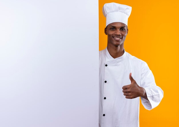 Joven cocinero afroamericano sonriente en uniforme de chef se encuentra detrás de la pared blanca y los pulgares arriba aislados en la pared naranja