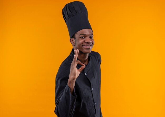 Joven cocinero afroamericano sonriente en uniforme de chef gestos ok signo de mano aislado en la pared naranja
