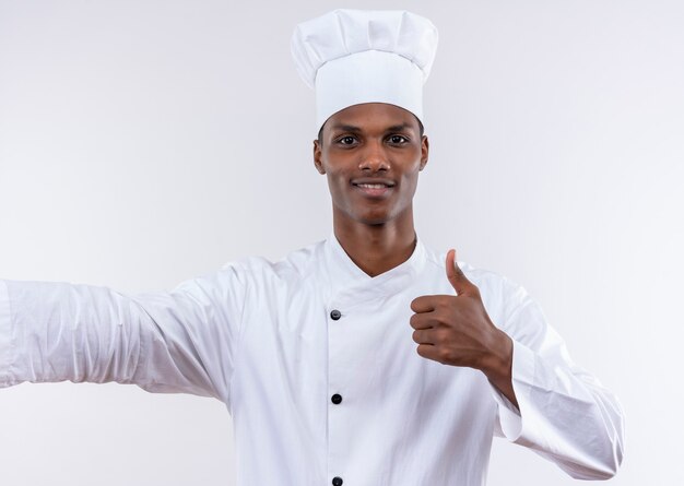 Joven cocinero afroamericano sonriente en uniforme de chef pulgares arriba aislado en la pared blanca