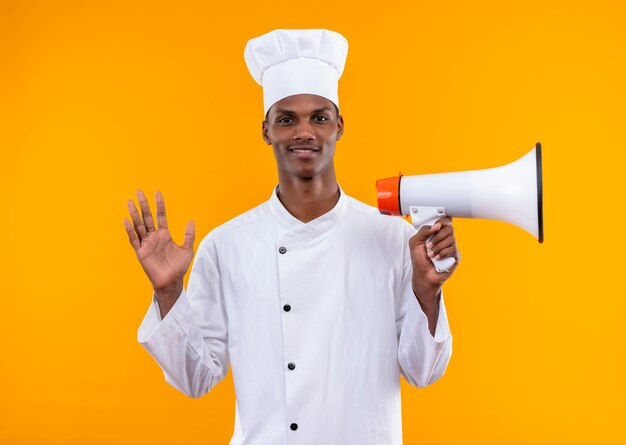 Joven cocinero afroamericano sonriente en uniforme de chef sostiene altavoz aislado en la pared naranja