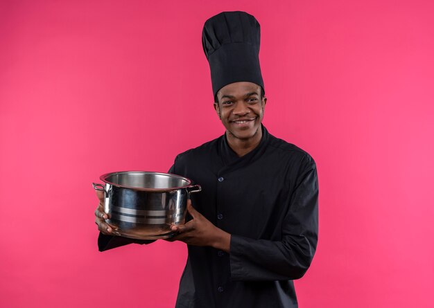 Joven cocinero afroamericano sonriente en uniforme de chef sostiene una cacerola aislada en la pared rosa