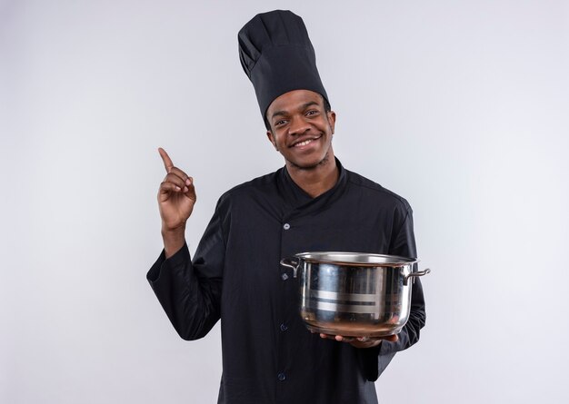 Joven cocinero afroamericano sonriente en uniforme de chef sostiene una cacerola y apunta hacia arriba aislado en la pared blanca