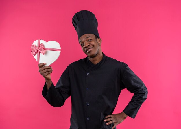 Joven cocinero afroamericano sonriente en uniforme de chef sostiene una caja en forma de corazón aislada en la pared rosa