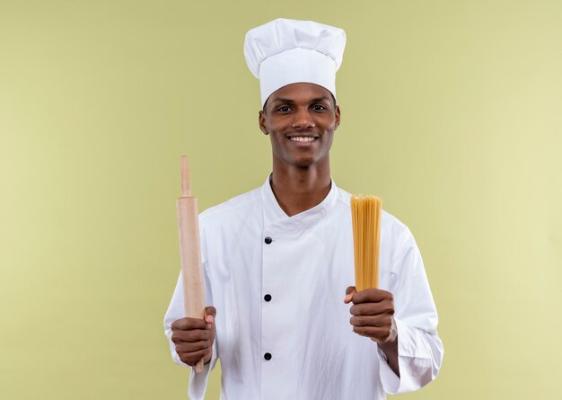 Joven cocinero afroamericano sonriente en uniforme de chef sostiene un rodillo y un montón de espaguetis en manos aisladas en la pared verde