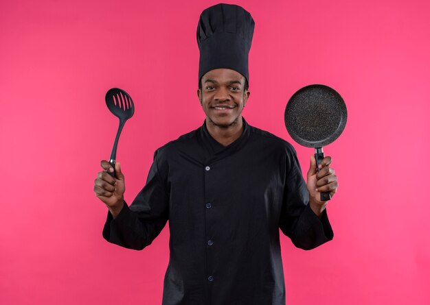 Joven cocinero afroamericano sonriente en uniforme de chef sostiene una sartén y una espátula aislada en la pared rosa