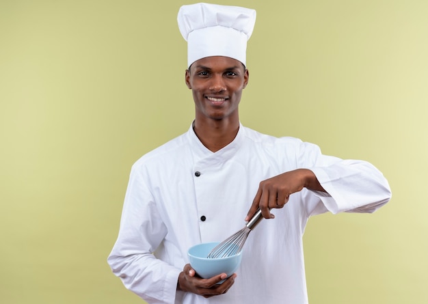 Joven cocinero afroamericano sonriente en uniforme de chef sostiene un tazón y un batidor aislado en la pared verde