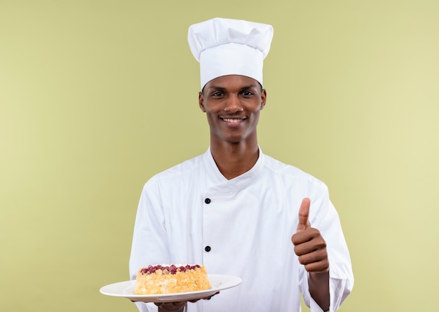 Joven cocinero afroamericano sonriente en uniforme de chef sostiene la torta en el plato y los pulgares hacia arriba aislados en la pared verde