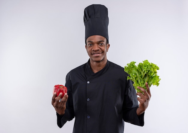 Joven cocinero afroamericano sonriente en uniforme de chef tiene pimiento rojo y ensalada verde aislado en la pared blanca