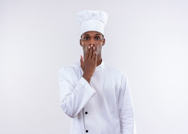 Joven cocinero afroamericano sorprendido en uniforme de chef pone la mano en la boca aislada en la pared blanca