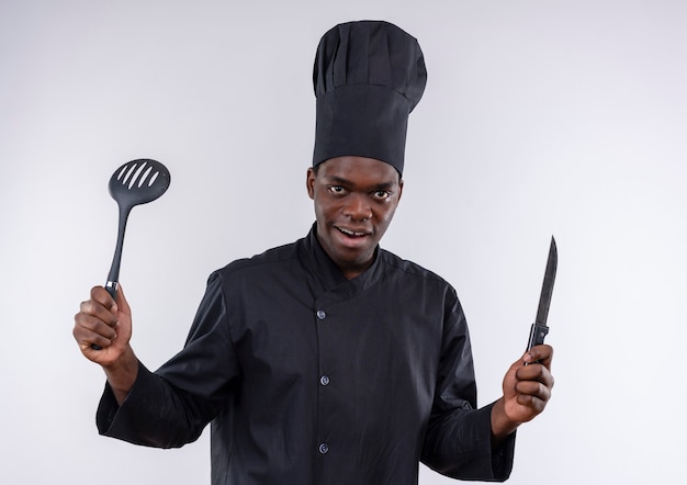 Joven cocinero afroamericano sorprendido en uniforme de chef sostiene cuchillo y espátula en blanco con espacio de copia