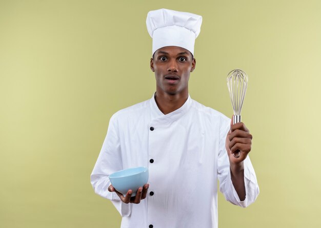 Joven cocinero afroamericano sorprendido en uniforme de chef sostiene un tazón y un batidor aislado en la pared verde