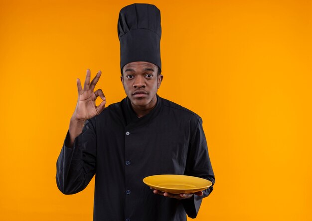 Joven cocinero afroamericano en uniforme de chef gestos ok signo de mano y sostiene placa aislada sobre fondo naranja con espacio de copia