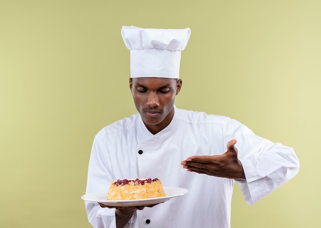 Joven cocinero afroamericano en uniforme de chef sostiene la torta en el plato y señala con la mano aislada sobre fondo verde con espacio de copia