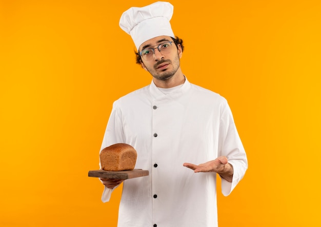 Joven cocinero enojado con uniforme de chef y gafas sosteniendo y apunta con la mano a la barba en la tabla de cortar aislada en la pared amarilla