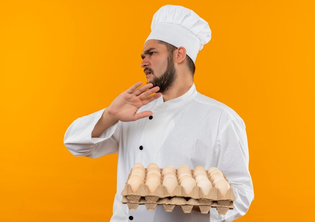 Joven cocinero enojado con uniforme de chef sosteniendo una caja de huevos mirando al lado y gesticulando no aislado en el espacio naranja