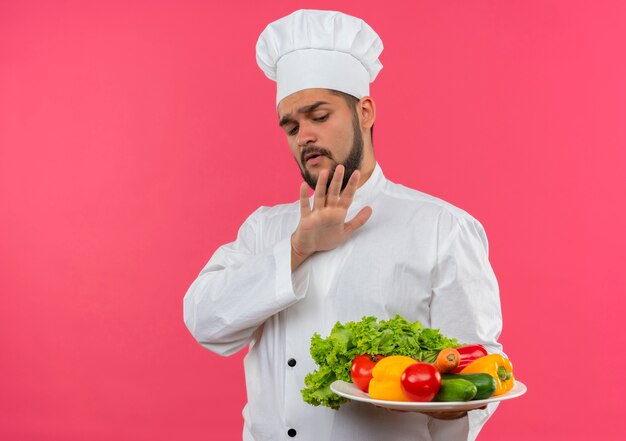 Joven cocinero enojado con uniforme de chef sosteniendo y mirando el plato de verduras y gesticulando no aislado en el espacio rosa