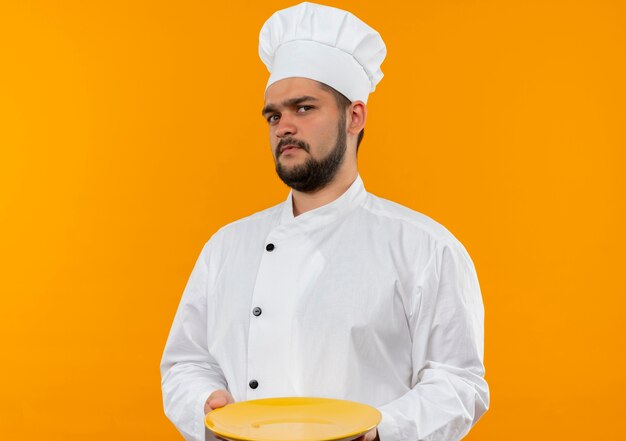 Joven cocinero enojado en uniforme de chef sosteniendo un plato vacío aislado en el espacio naranja