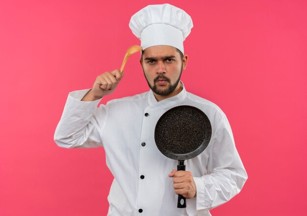 Joven cocinero enojado con uniforme de chef sosteniendo una sartén y tocando la cabeza con una cuchara aislada en el espacio rosa