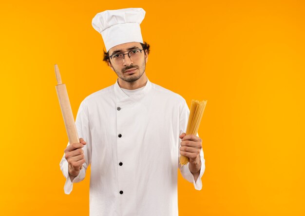 Joven cocinero enojado vistiendo uniforme de chef y gafas sosteniendo espaguetis y rodillo aislado en la pared amarilla