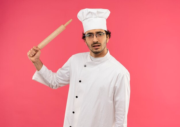 Joven cocinero enojado vistiendo uniforme de chef y gafas sosteniendo un rodillo alrededor del hombro