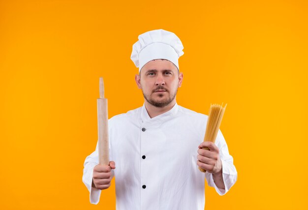Joven cocinero guapo en uniforme de chef con rodillo y pasta de espagueti mirando aislado en espacio naranja