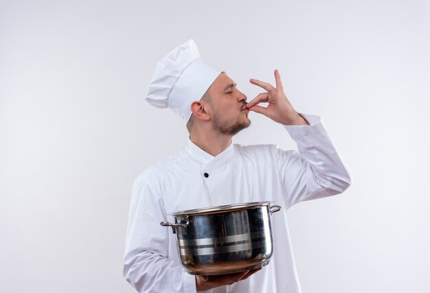 Joven cocinero guapo en uniforme de chef sosteniendo la caldera y haciendo un gesto sabroso en un espacio en blanco aislado