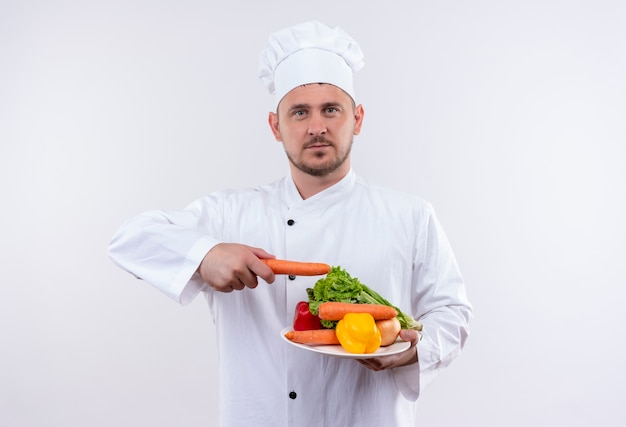 Joven cocinero guapo en uniforme de chef sosteniendo un plato con verduras y apuntando a ellos con zanahoria mirando en espacio en blanco aislado