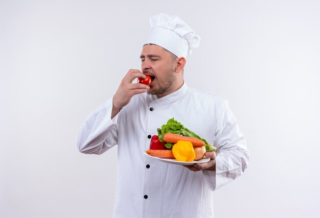 Joven cocinero guapo en uniforme de chef sosteniendo un plato con verduras y mordiendo tomate con los ojos cerrados en un espacio en blanco aislado