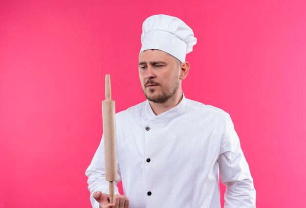 Joven cocinero guapo en uniforme de chef sosteniendo un rodillo mirándolo aislado en el espacio rosa