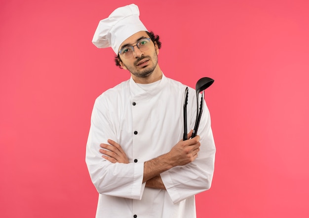 Joven cocinero con uniforme de chef y gafas sosteniendo una espátula y cruzando las manos