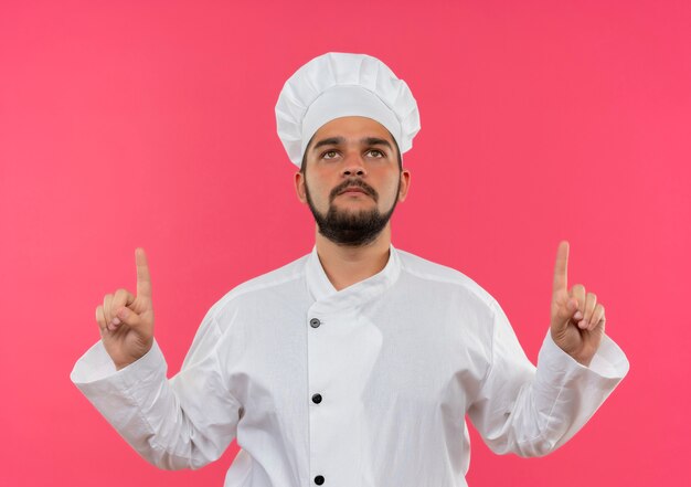 Joven cocinero en uniforme de chef mirando y apuntando hacia arriba aislado en el espacio rosa