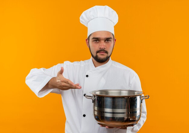 Joven cocinero en uniforme de chef sosteniendo y apuntando con la mano a la olla y mirando aislado en el espacio naranja