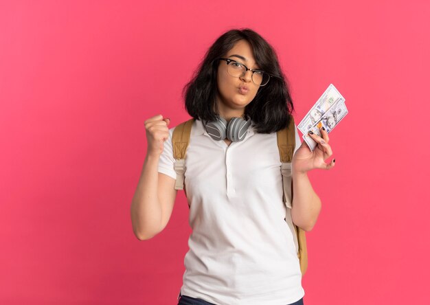 Joven colegiala bastante caucásica alegre con auriculares en el cuello con gafas y bolsa trasera tiene dinero y levanta la mano en rosa con espacio de copia