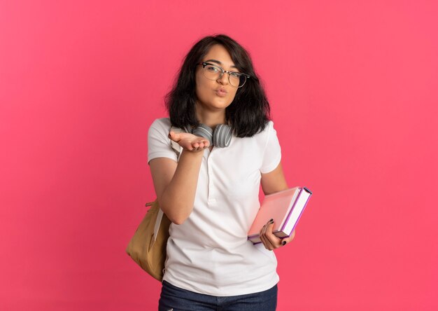 Joven colegiala bastante caucásica alegre con gafas y bolso trasero con auriculares en el cuello envía beso con la mano sosteniendo libros en rosa con espacio de copia