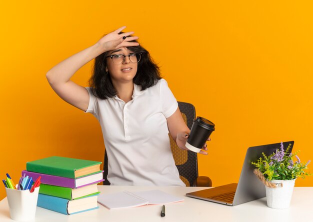 Joven colegiala bastante caucásica ansiosa con gafas se sienta en el escritorio con herramientas escolares pone la mano en la frente sosteniendo la taza de café en naranja con espacio de copia