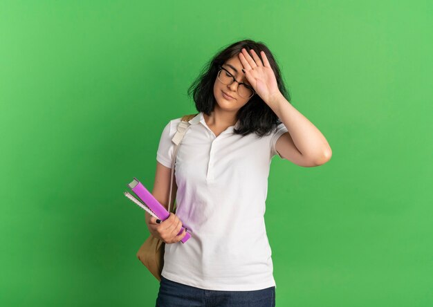 Joven colegiala bastante caucásica cansada con gafas y bolso trasero pone la mano en la frente sosteniendo libros en verde con espacio de copia