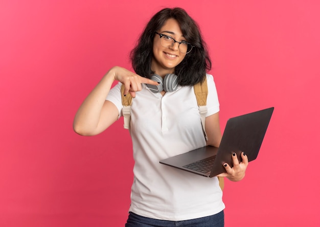 Joven colegiala bastante caucásica complacida con auriculares en el cuello con gafas y bolsa trasera sostiene y apunta a la computadora portátil en rosa con espacio de copia