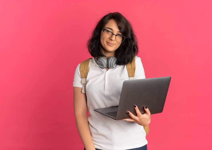 Foto gratuito joven colegiala bastante caucásica complacida con auriculares en el cuello con gafas y bolsa trasera sostiene laptop mirando a cámara en rosa con espacio de copia
