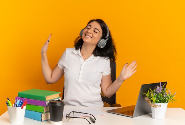 Joven colegiala bastante caucásica complacida en auriculares se sienta en el escritorio con herramientas escolares con las manos levantadas aisladas en el espacio naranja con espacio de copia