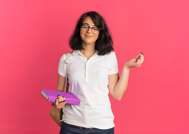 Joven colegiala bastante caucásica complacida con gafas y bolsa trasera sostiene bolígrafo y libro y levanta la mano con los ojos cerrados en rosa con espacio de copia