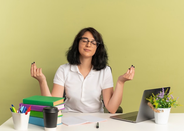 Foto gratuita joven colegiala bastante caucásica complacida con gafas se sienta en el escritorio con herramientas escolares finge meditar con las manos levantadas aisladas en el espacio verde con espacio de copia