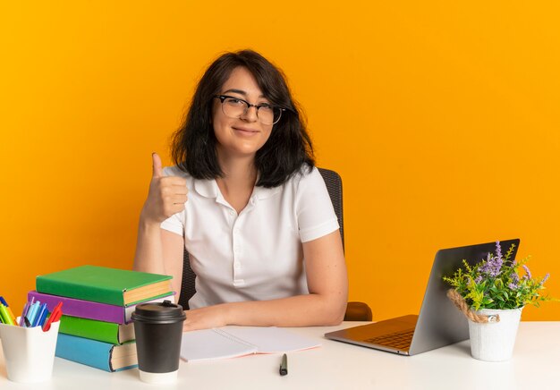 Joven colegiala bastante caucásica complacida con gafas se sienta en un escritorio con herramientas escolares Thumbs up en naranja con espacio de copia