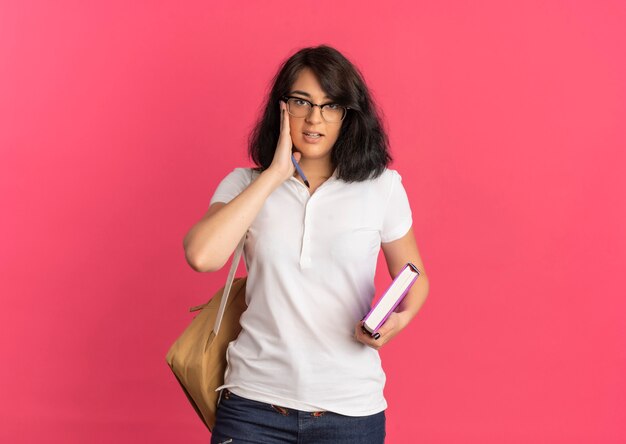 Joven colegiala bastante caucásica confiada con gafas y bolso trasero pone la mano en la cara sosteniendo la pluma y el libro en rosa con espacio de copia